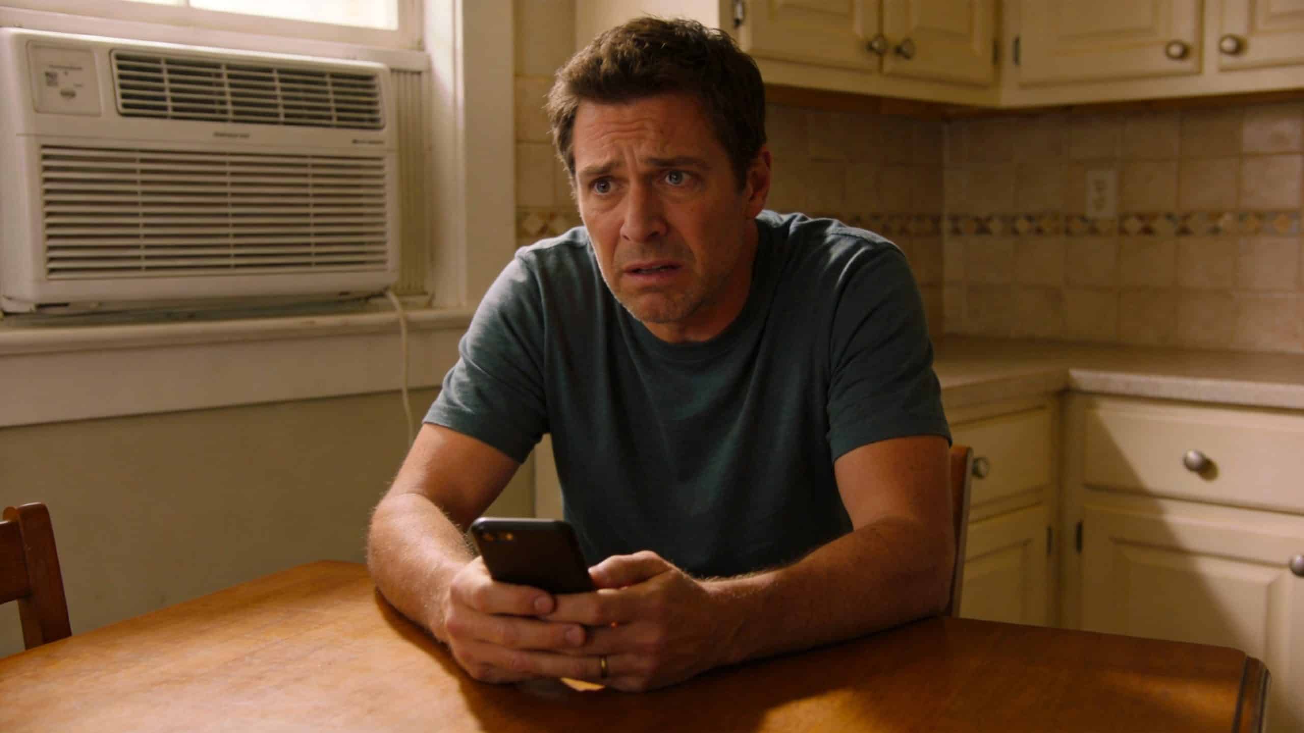 Photorealistic 16:9 photo of a homeowner at a kitchen table looking worried at a smartphone (high-converting AC repair page)